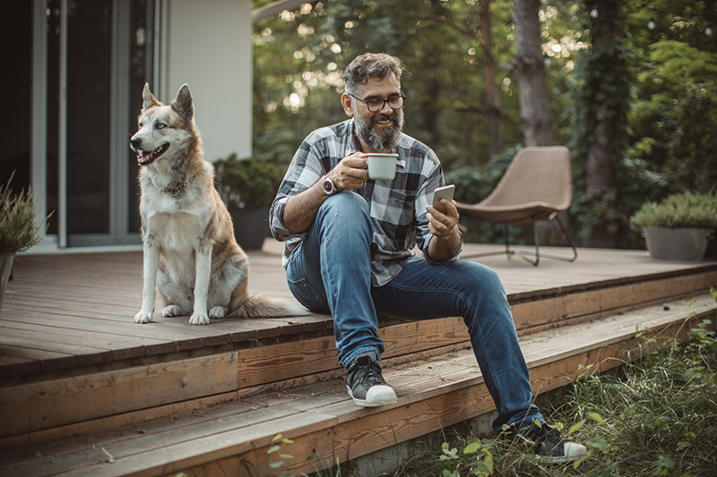 Mature men at his cottage resting on pprch with his dog. Sitting in cahair, drinking coffee and using smart phone. Wearing casual clothing.